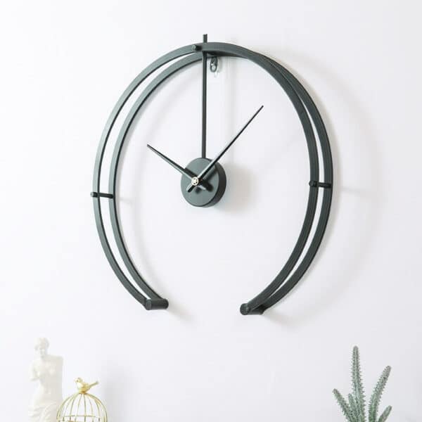 Horloge murale noire en arc de cercle en métal installée sur un mur blanc, avec une petite plante posée en bas à droite