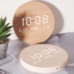 Horloge murale en bois ronde et digitale posé sur une table en marbre et qui reflète l'horloge, sur la gauche un petit pot avec un petite plante verte dedans