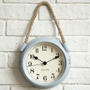 Horloge classique avec cadran blanc , aiguilles noires, et cadre bleu clair, elle est suspendue par une corde sur un mur en brique blanche