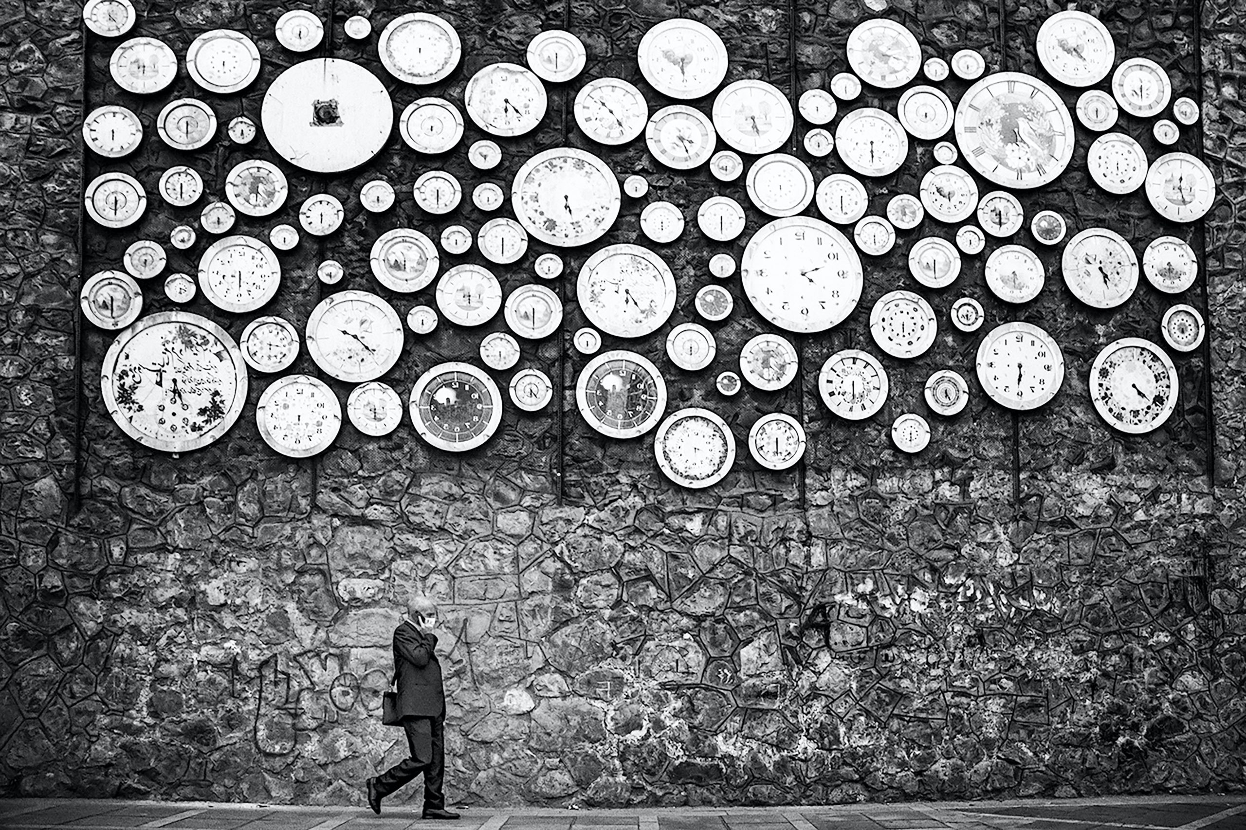 Personne qui marche dans la rue en longeant un mur en pierre sur lequel se trouve sur toute la partie haute des dizaine d'horloges murales rondes instalées les unes à côté des autres