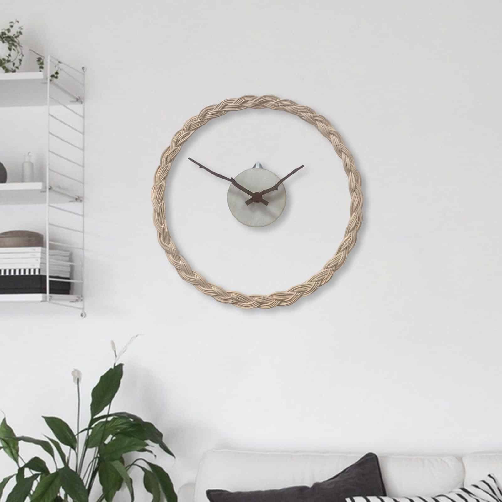 Horloge transparente avec le tour en rotin et le centre metal avec les aiguilles style bois, installée sur un mur blanc dans un salon avec des étagères à gauche et en bas des plantes