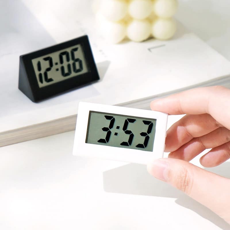 Mini horloge digitale de table, l'une est noire posée sur un support blanc et une autre blanche est tenue par une main