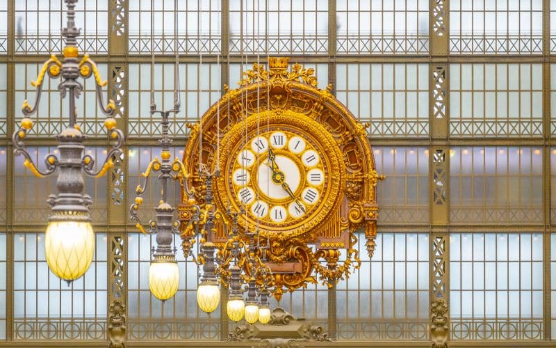 Grande horloge dorée avec de nombreuses moulures et des chiffres romains. Elle semble être accrochée dans une gare ou un grand monument.