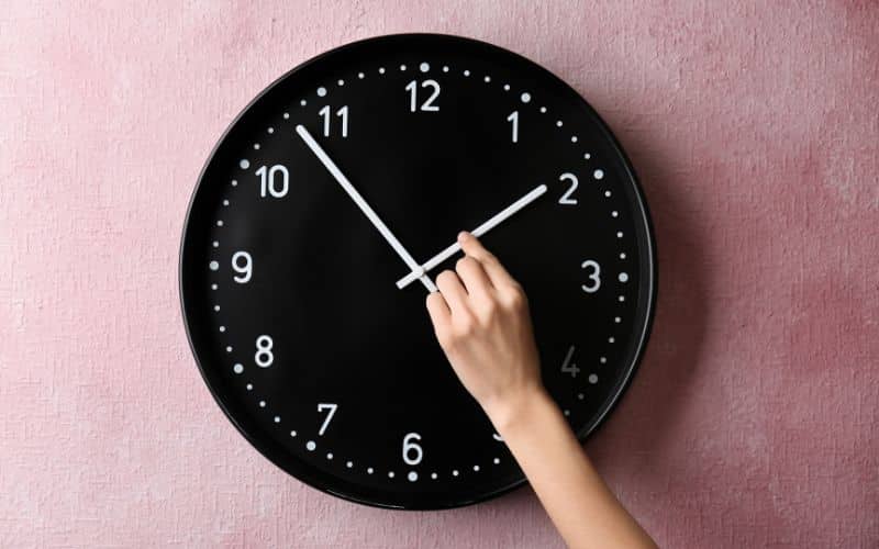 Horloge murale noire, accrochée sur un mur rose, une main déplace l'aiguille des heures.